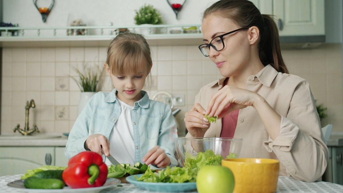 Cute loving daughter is helping mother with salad cutting cucumbers feeding young woman and laughing having fun at home. People and cookery concept.